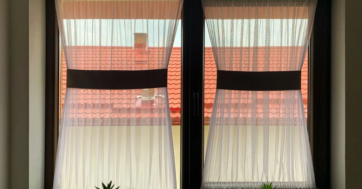 Une fenêtre avec des rideaux blancs transparents, vue sur un toit rouge, et de petites plantes en pot sur le rebord.