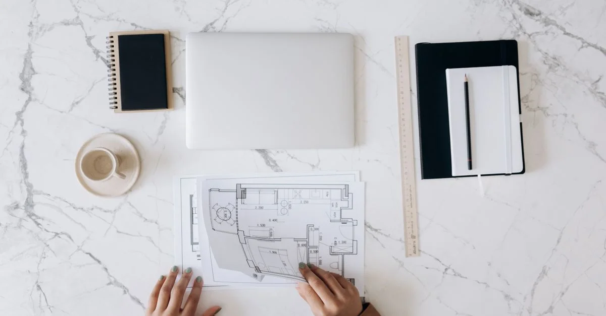 Vue de dessus du bureau d'un architecte présentant des plans, un ordinateur portable, un carnet et une tasse de café sur une surface en marbre.