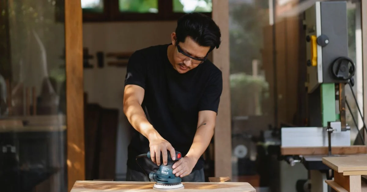Un menuisier concentré portant des lunettes de sécurité utilise une ponceuse pour façonner une planche en bois dans un atelier bien éclairé.